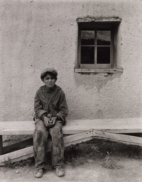 A black-and-white photograph of a squinting medium-skinned boy wearing a cap, jacket, and pants with patches on the knees, seated on a wooden bench in front of an adobe building.