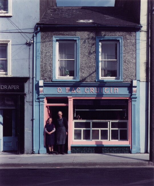 A color photograph of a White man and woman standing in the doorway of a small, two-story storefront.