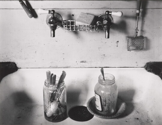 A black-and-white photograph of a two-tap sink with two dirty mason jars full of spoons.