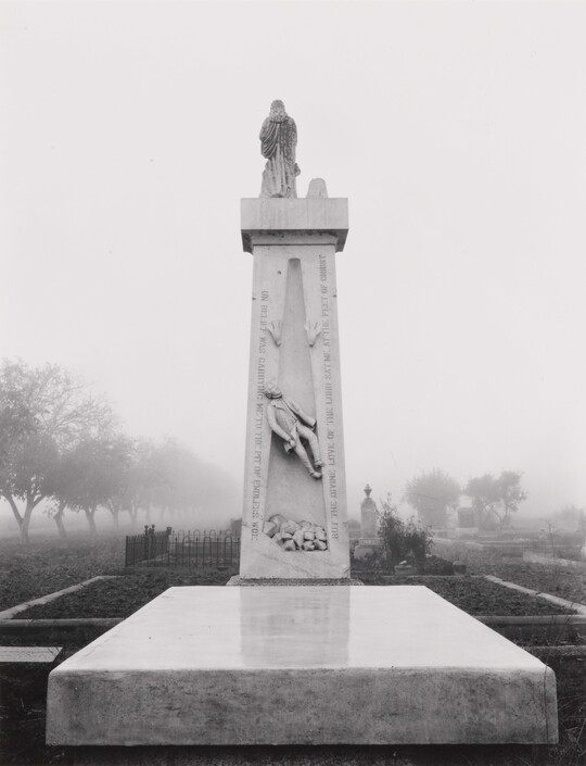 A black-and-white photograph of a tall, vertical stone monument rising behind a flat stone slab in a cemetery.