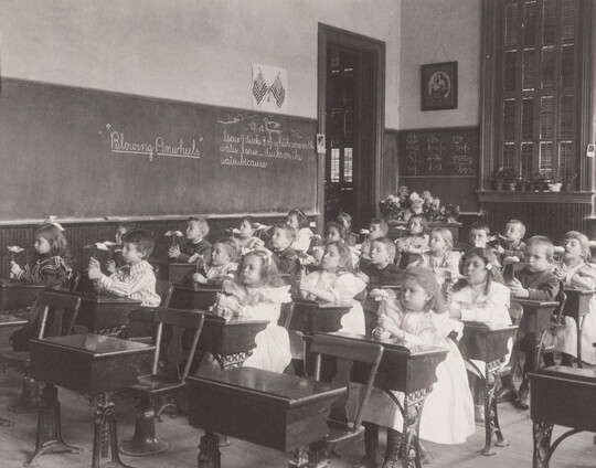 A black-and-white photograph of a classroom of young children seated at desks blowing on pinwheels.