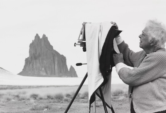 A black-and-white photograph of an older White woman with a box camera on a tripod and a tall rock formation in the background.