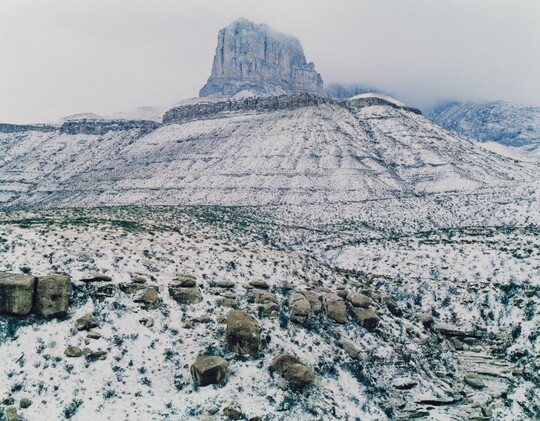 A color photograph of a snowy landscape featuring a tall butte and distant mountains obscured by snowy haze.
