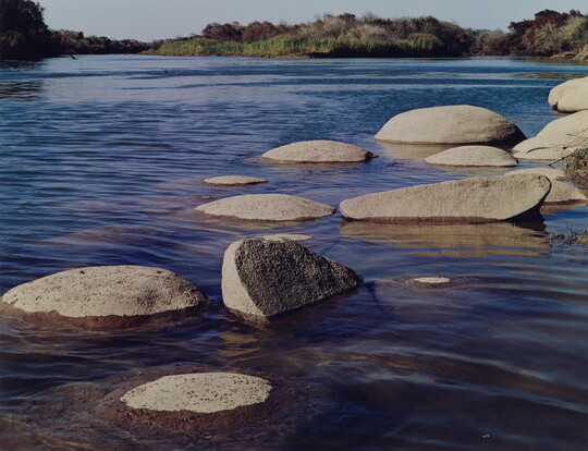 A color photograph of many, large smooth boulders in a river reflecting a blue sky.