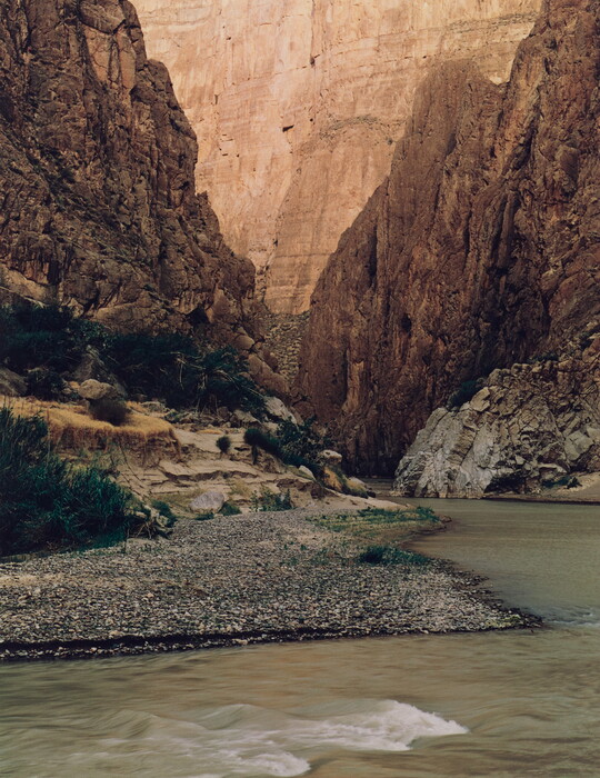 A color photograph of a river surrounded by tall rocky cliffs with some green vegetation.
