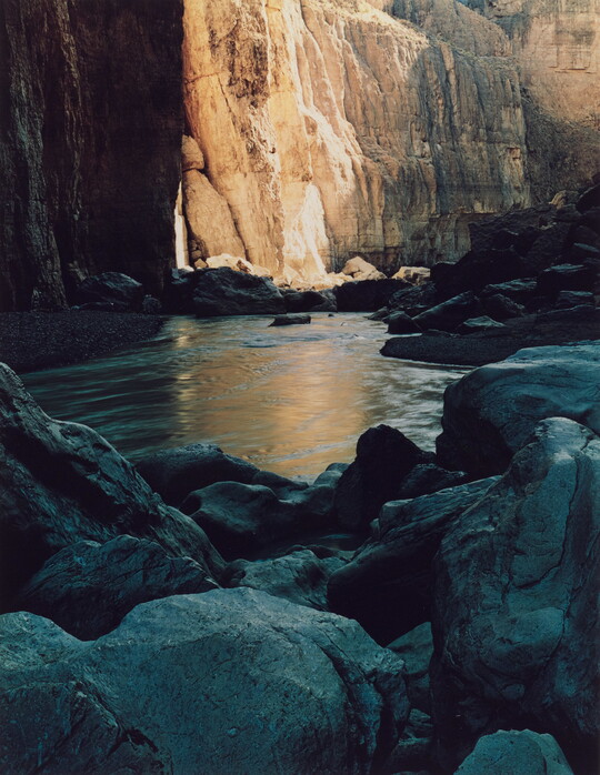 A color photograph of large boulders resting in still water at the base of a cliff.
