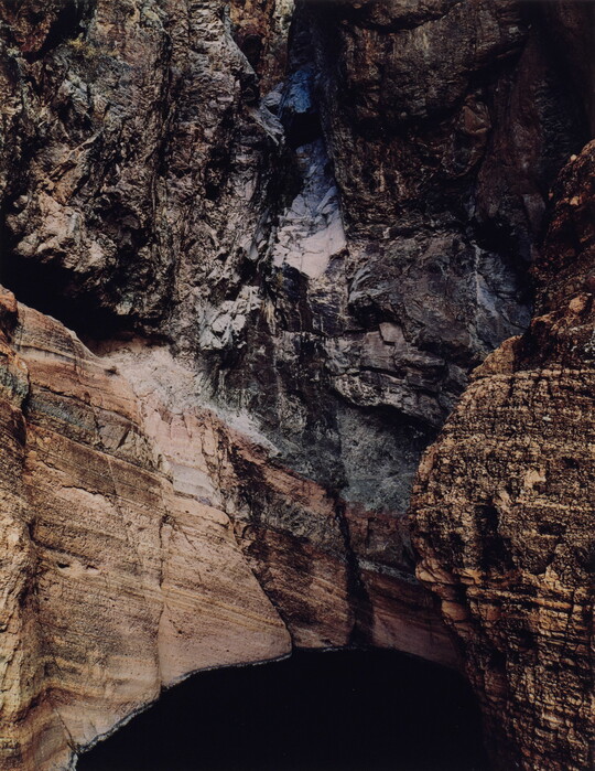 A color photograph of the side of a rocky cliff showing layers of rock above a dark hole.