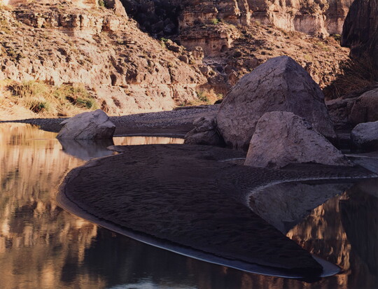 A color photograph of large boulders on a sandy riverside beach and a rocky cliff in the background.