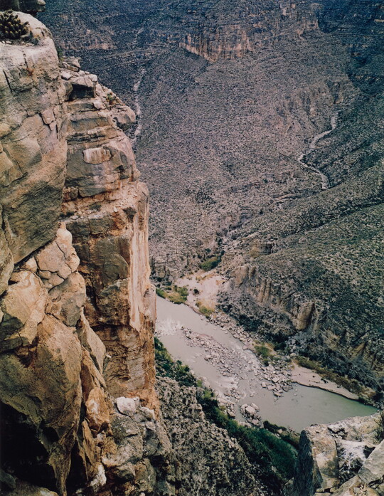 A color photograph taken from the top of a cliff overlooking a river surrounded by other rocky cliffs.