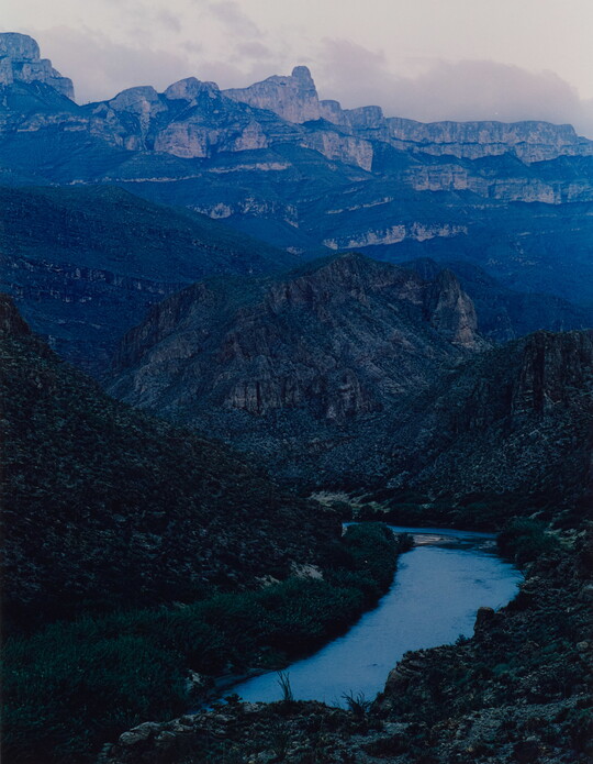 A color photograph of a river in a valley surrounded by blue mountains and cliffs.