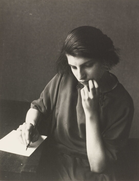 A black-and-white photograph of a young White woman seated at a table writing on a piece of paper.
