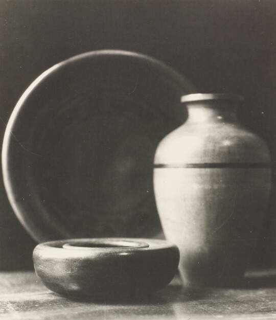 A black-and-white photograph of a short wood bowl, a large pear-shaped vase, and a standing plate behind them.