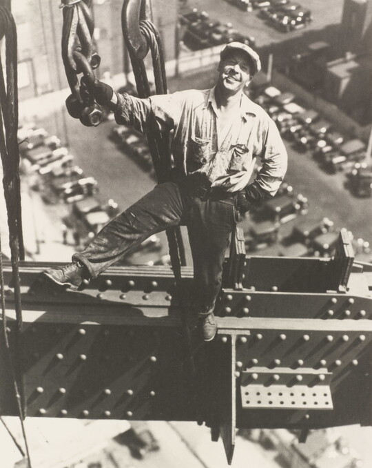 A black-and-white photograph of a smiling White man standing on a beam holding onto a chain, wearing a newsboy cap, cars in parking lots far below.