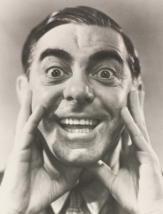 A black-and-white portrait photograph of a White man, hair slicked back, hands framing his cheeks, mouth open in a wide smile.