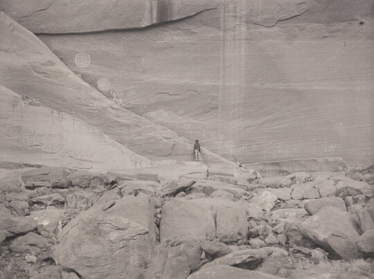 A black-and-white photograph of a person standing against a large cliff face, and petroglyphs high on the cliff wall.