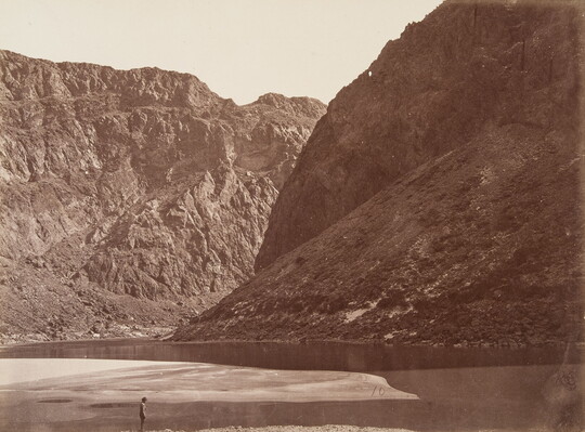 A black-and-white photograph of a person standing in a river surrounded by large rocky mountains.