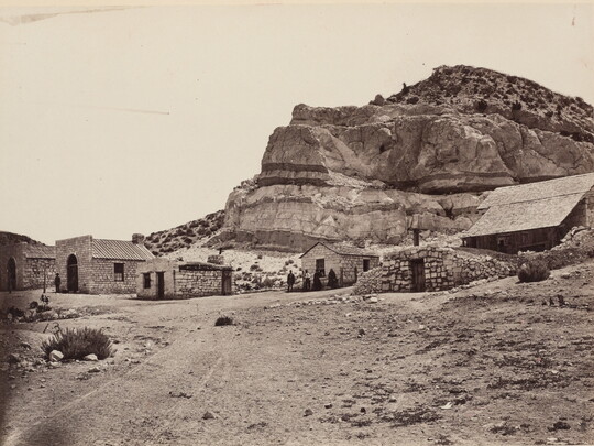 A black-and-white photograph of a group of small stone buildings at the base of a cliff in a desert landscape.