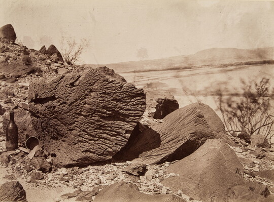 A black-and-white photograph of a striated rock and a glass bottle standing next to it in a desert landscape.