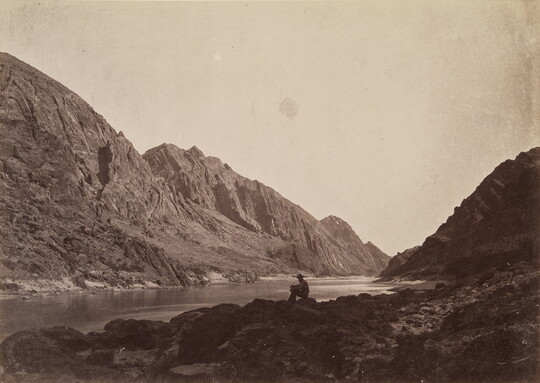A black-and-white photograph of a person sitting on a rock next to a calm river surrounded by steep cliffs and mountains.