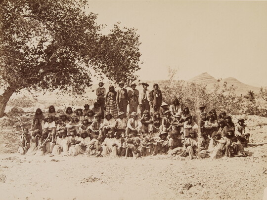 A black-and-white photograph of a large group of Indigenous people, some seated and some standing, under a tree with mountains in the distance.