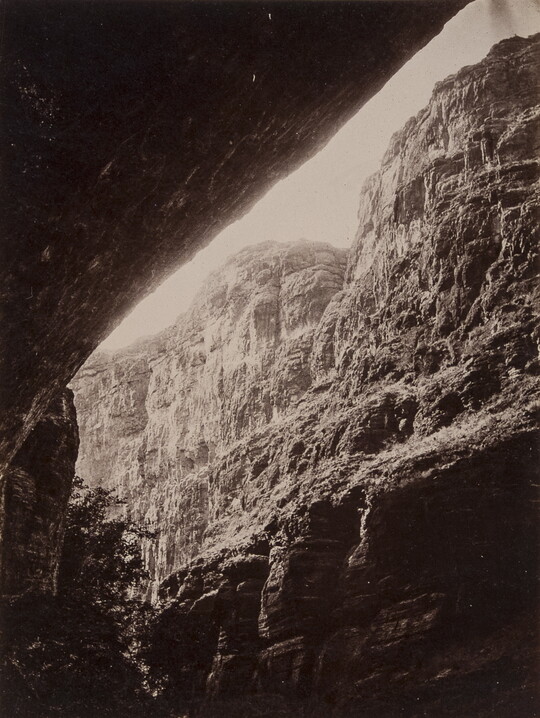 A black-and-white photograph of a steep cliff face from underneath a rock overhang.