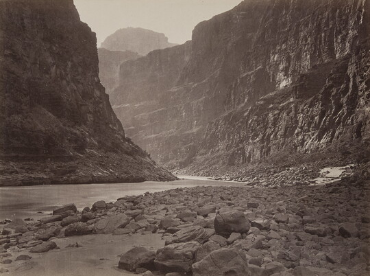A black-and-white photograph of a calm river at the rocky bottom of a canyon.