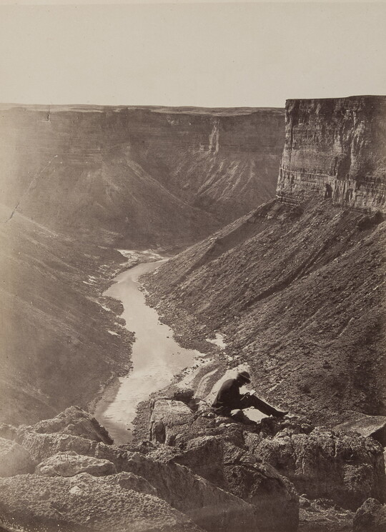 A black-and-white photograph of a person sitting on rocks above a river running through a canyon surrounded by tall cliffs.