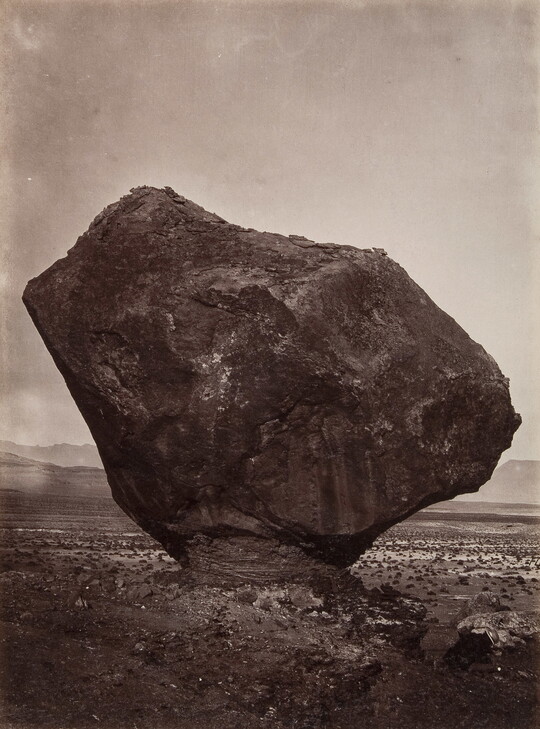 A black-and-white photograph of a large rock formation balanced on a narrow pedestal in a desert.