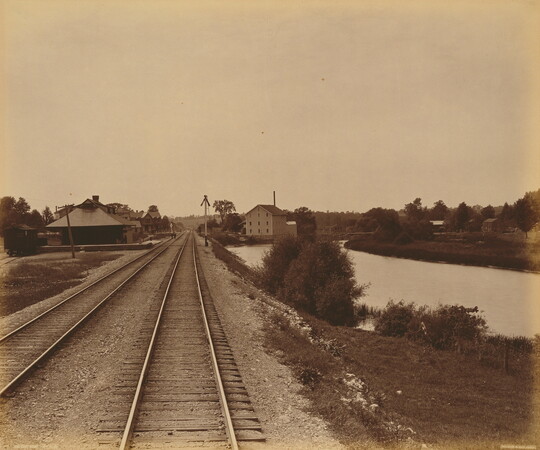 A sepia-toned photograph of railroad tracks leading to several buildings, and water to the right of the tracks.