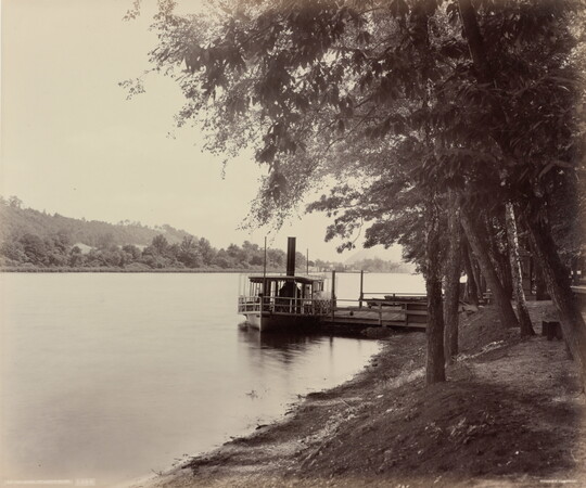 A black-and-white photograph of a boat docked on a river and trees on the shore.