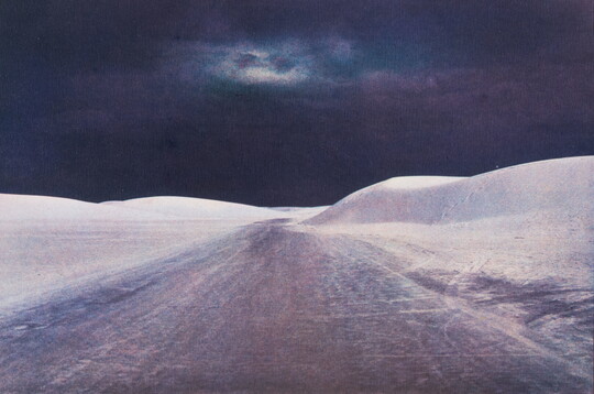 A color photograph of rolling white sand dunes on either side of a road under a dark and cloudy night sky.