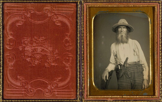 A black-and-white portrait photograph, in a gold frame and red velvet case, of a bearded White man wearing a hat, white shirt, and plaid pants with a knife tucked into the waistband.