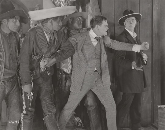 A black-and-white photograph of three medium-skinned men wearing sombreros and one White man in a suit restraining an angry White man in a three-piece suit.