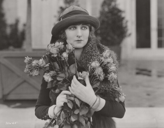 A black-and-white photograph of a White woman wearing a bucket hat and clutching a large bouquet of long-stemmed roses to her chest.