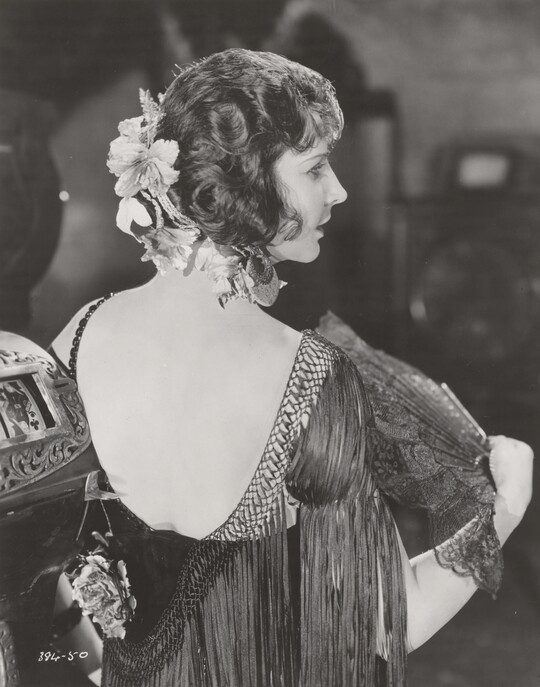 A black-and-white photograph, taken from the back, of a White woman wearing a low-back fringed frock, and fluttering a lacy fan.