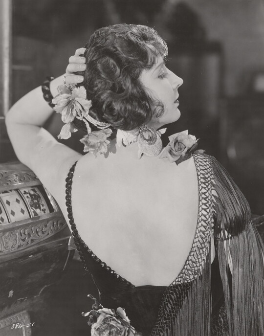A black-and-white photograph, taken from the back, of a White woman, flowers in her hair, wearing a low-back fringed frock.