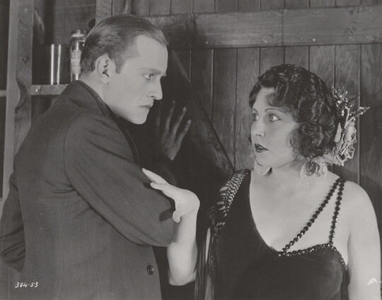 A black-and-white photograph of a White man looking intently at a White woman; she has her hand on his arm and looks behind him.