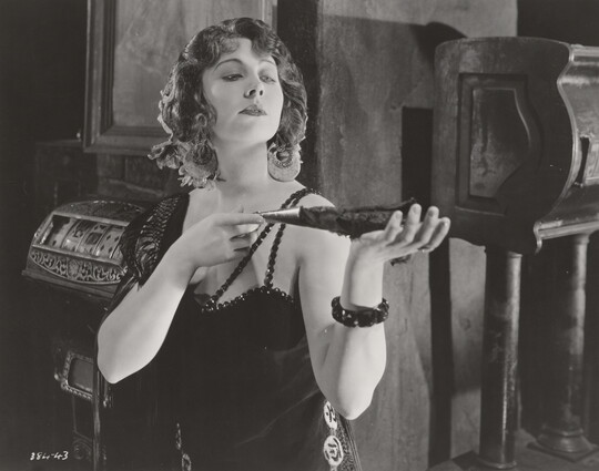 A black-and-white photograph of a White woman wearing a 1920s-style dress holding a feathered fan in her outstretched hands.