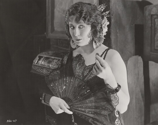 A black-and-white photograph of a White woman wearing a 1920s-style dress holding an open fan in one hand and a coin in the other.