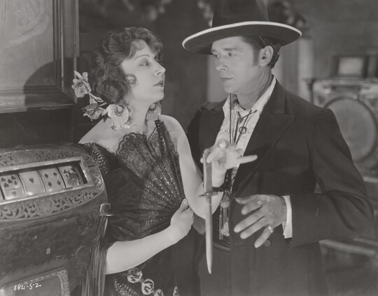 A black-and-white photograph of a White woman wearing a 1920s-style dress holding an open fan against her chest and a dagger as the White man next to her reaches for the dagger.