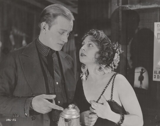 A black-and-white photograph of a White man and woman dressed in 1920s attire looking at each other as the man points to a coffee pot he is holding.