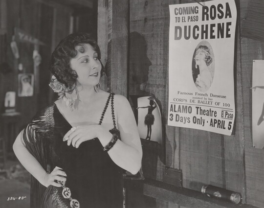 A black-and-white photograph of a White woman wearing a 1920s-style dress, one hand on her hip, looking at a poster nailed to a wall.