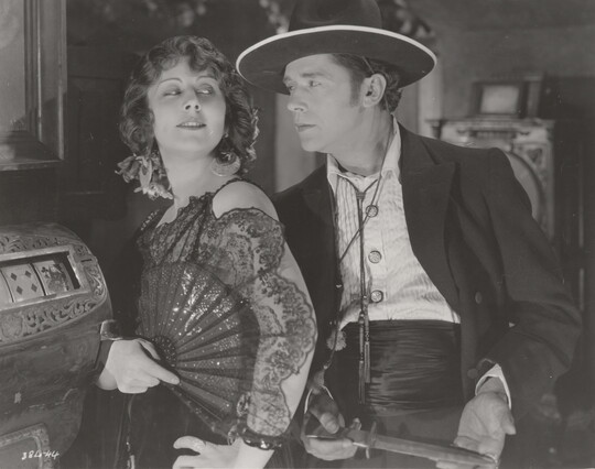 A black-and-white photograph of a White woman wearing a 1920s-style dress holding an open fan against her shoulder as the White man behind her holds a dagger.