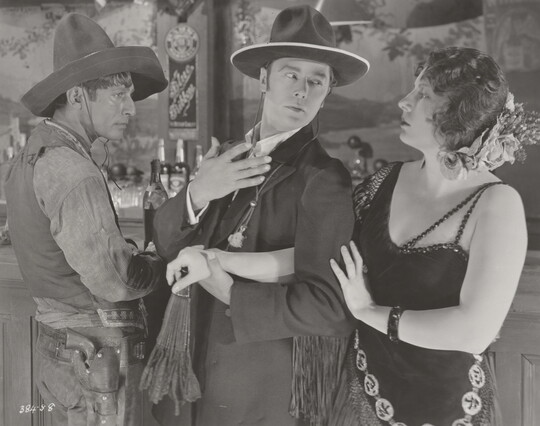 A black-and-white photograph of a medium-skinned man wearing a sombrero, a White man wearing a suit, and a White woman wearing a 1920s-style dress at a bar.