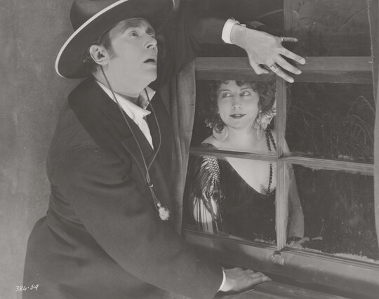 A black-and-white photograph of a White man standing next to a window, one hand under the window frame, while a White woman wearing a 1920s-style dress looks at him from the other side of the window.