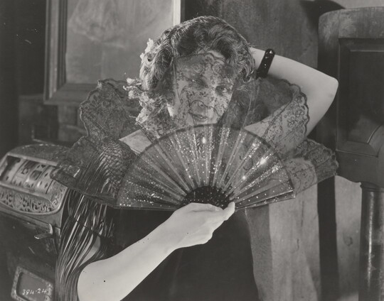A black-and-white photograph of a White woman holding an open lace fan in front of her face.