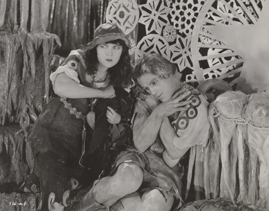 A black-and-white photograph of a White woman and man dressed in theatrical costumes in front of a set depicting snowflakes and icicles.