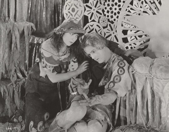 A black-and-white photograph of a White woman and man dressed in theatrical costumes in front of a set depicting snowflakes and icicles.
