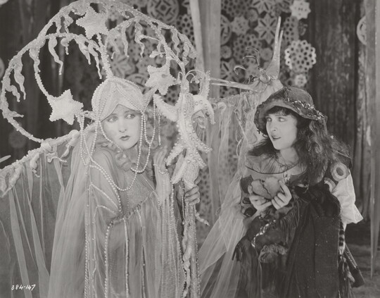 A black-and-white photograph of two White women in theatrical costumes in front of a set depicting snowflakes and icicles.