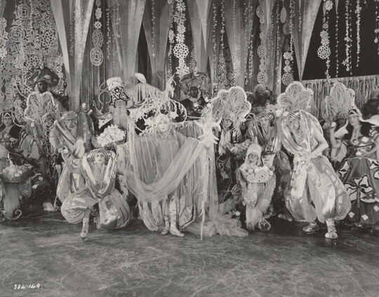 A black-and-white photograph of a group of people on a stage wearing elaborate costumes in front of a set depicting snowflakes and icicles.
